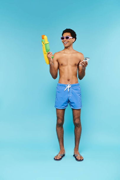 Şık güneş gözlüklü neşeli Afrikalı Amerikalı adam elinde telefon ve su tabancasıyla mavi arka planda. - Fotoğraf, Görsel