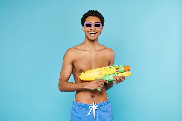 jovem homem americano africano alegre em calções de banho com óculos de sol elegantes posando com arma de água - Foto, Imagem