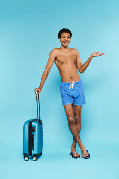 alegre joven afroamericano en traje de baño posando junto a la maleta y sonriendo a la cámara - Foto, Imagen
