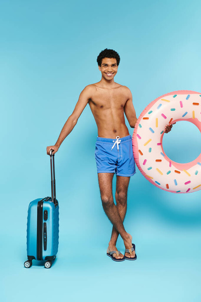 cheerful african american man in swimming trunks with swimming ring and suitcase smiling at camera - Photo, Image