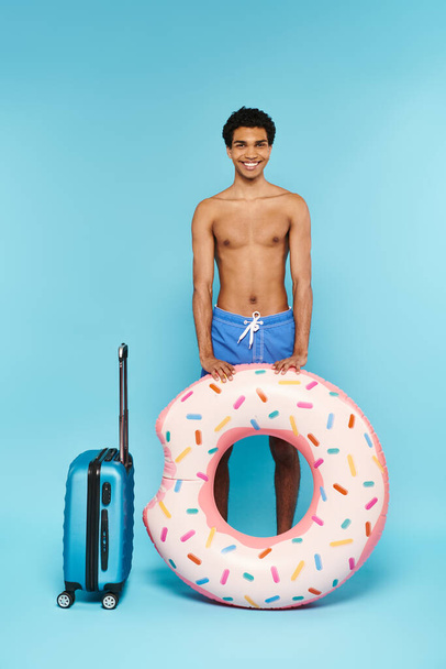 radosny Afrykański Amerykanin w kąpielówkach z pierścieniem kąpielowym i walizką uśmiechnięty do kamery - Zdjęcie, obraz