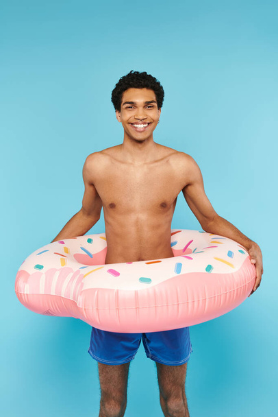 handsome cheerful african american man with swimming ring on blue backdrop looking at camera - Photo, Image