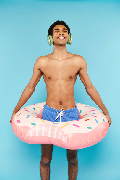 fröhlicher afrikanisch-amerikanischer Mann in Badehose mit aufblasbarem Donut und Musik im Kopfhörer - Foto, Bild
