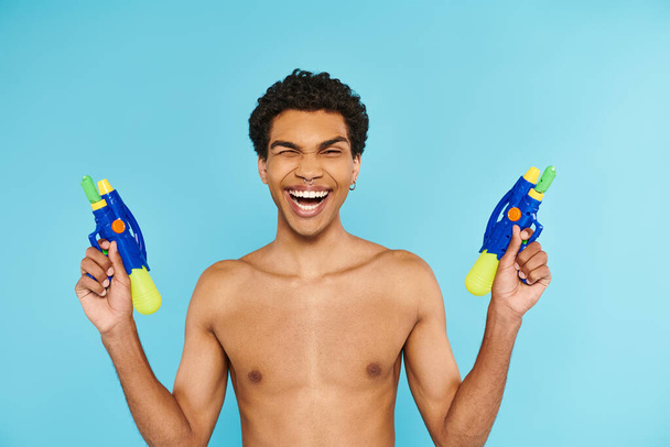 positieve knappe Afro-Amerikaanse man poseren met twee waterpistolen en glimlachen naar de camera gelukkig - Foto, afbeelding