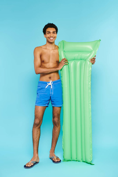 alegre afroamericano hombre en traje de baño posando con colchón de aire y sonriendo a la cámara - Foto, Imagen