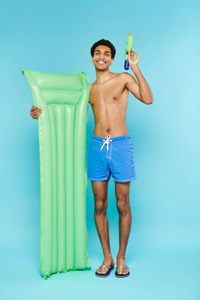 pozitív fiatal afro-amerikai férfi pózol levegő matrac és vízipisztoly kék háttér - Fotó, kép