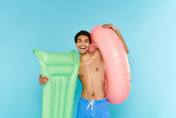 alegre afroamericano hombre posando con colchón de aire y donut inflable y sonriendo a la cámara - Foto, imagen