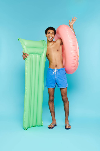 alegre afroamericano hombre posando con colchón de aire y donut inflable y sonriendo a la cámara - Foto, Imagen