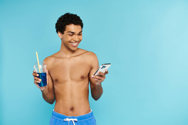 Radosny, afrykański Amerykanin w kąpielówkach, patrzący na telefon i trzymający koktajl w dłoni. - Zdjęcie, obraz