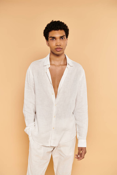привабливий молодий афроамериканський чоловік у білому вбранні позує на бежевому тлі і дивиться на камеру - Фото, зображення