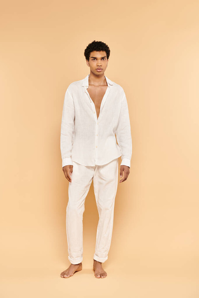 alluring young african american man in white attire posing on beige backdrop and looking at camera - Photo, Image