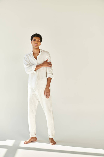 елегантний афроамериканський чоловік у білому вбранні позує босоніж і дивиться на камеру на бежевому тлі - Фото, зображення