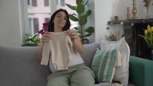 Anyai előrelátás - Várandós anya késői Terhesség Megmutatja a baba ruhák a kamera, Kényelmesen ül a kanapén, átölelve az örömök a közelgő anyaság - Felvétel, videó