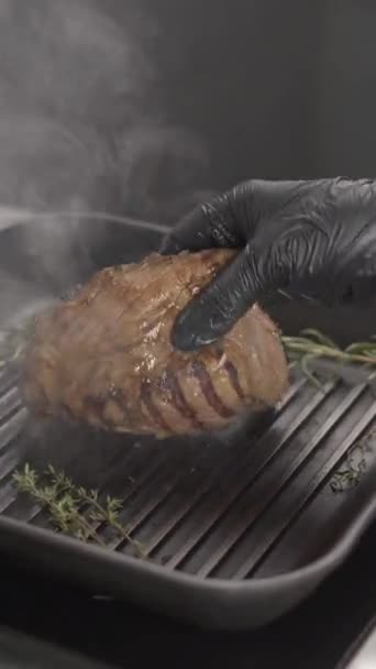 Close-up piece of steak is fried on the grill pan in restaurant. Chef flips tasty grilled meat steak with rosemary in kitchen. Vertical view - Footage, Video