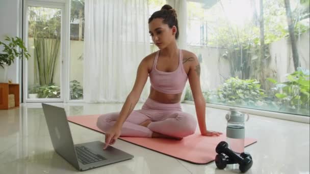 Amplio plano de joven gen Z mujer en ropa deportiva meditando en pose de loto delante del portátil mientras hace yoga en casa - Imágenes, Vídeo