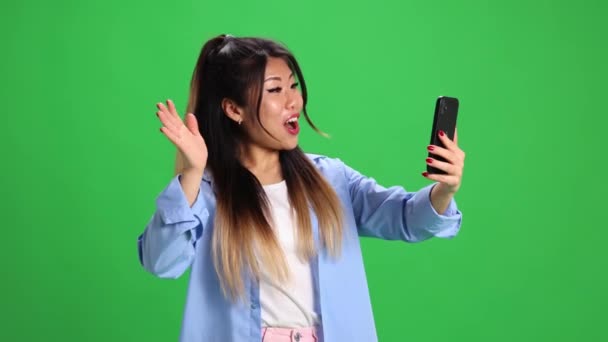 Uśmiechnięta Azjatka machająca witaniem podczas spotkania wideo w cyfrowym smartfonie na żywym zielonym tle studio. Pojęcie biznesu, nauki i pracy, start-up, sprzedaż, zakupy. - Materiał filmowy, wideo