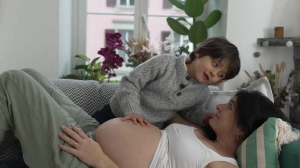 Sydämellinen hetki lapsen nojaten raskaana olevaan äitiin, kun hän lepää sohvalla, kaunis lämmin äidin ilo raskauden loppuvaiheessa, odottaen pikkuveljen saapumista. - Materiaali, video