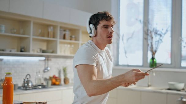 Un tipo escuchando música en la cocina usando auriculares inalámbricos de cerca. Hombre relajado alegre bailando en auriculares preparando sabrosos brindis para el desayuno en el acogedor interior de la cocina. Cocina melómana en auriculares - Foto, Imagen