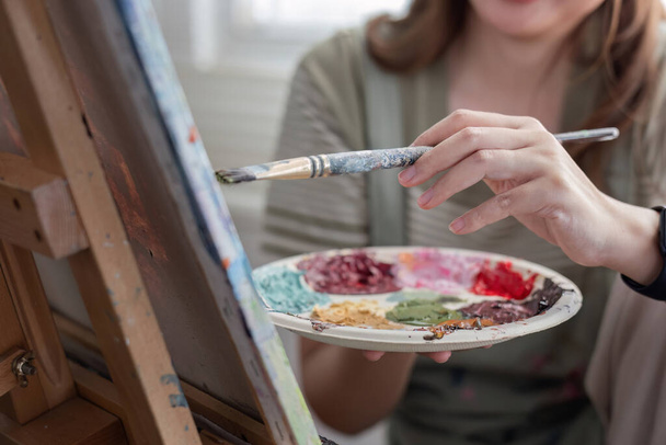 Молодая азиатская художница, держащая в руках палитру красочных красок, намеренно рисует акриловые краски на холсте в студии живописи.. - Фото, изображение