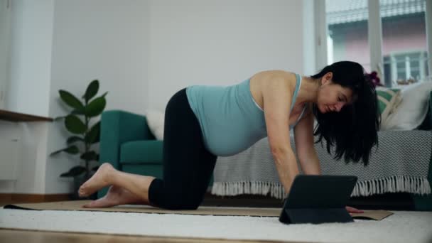 30s Вагітна жінка практикує йогу вдома - після цифрового тренера на планшеті для пренатальної рутини, підняття ноги на підлозі для спортивних вправ - Кадри, відео