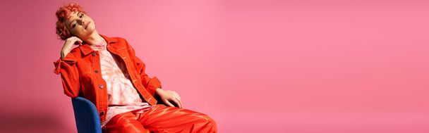 Erikoislaatuinen nainen istuu rauhallisesti sinisellä tuolilla elävän vaaleanpunaisen seinän edessä.. - Valokuva, kuva