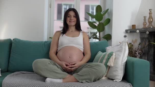 Portret szczęśliwej kobiety w ciąży siedzącej na kanapie w domu uśmiechniętej w późnym stadium ciąży, trzeci trymestr ciąży - Materiał filmowy, wideo