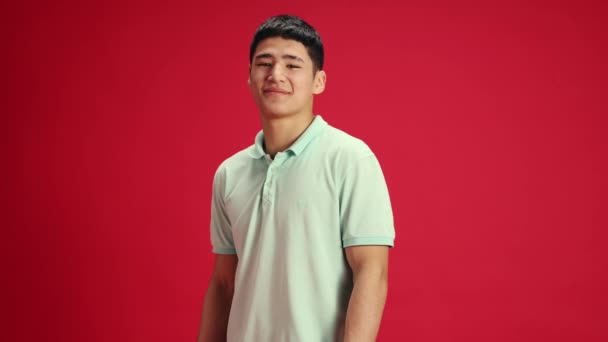 Jovem, bonito homem asiático vestindo roupas casuais com sorriso olhando para a câmera contra o fundo do estúdio vermelho. Conceito de emoções humanas, moda e beleza, auto-expressão, trabalho e hobby. Anúncio - Filmagem, Vídeo