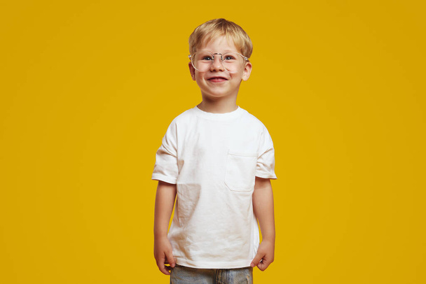 Vrolijke kleine slimme schooljongen met wit t-shirt en stijlvolle brillen, glimlachend en wegkijkend terwijl hij geïsoleerd over een gele achtergrond staat. Terug naar schoolconcept - Foto, afbeelding