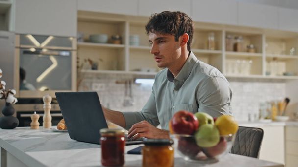 Businessman working at kitchen table with modern laptop close up. Serious young remote worker looking on computer screen making notes at notepad. Focused guy student studying online with notebook. - Photo, Image