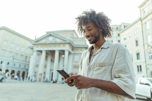 Мужчина с вьющимися волосами смотрит на свой телефон на городской площади. Сцена оживленная и оживленная, вокруг гуляют люди - Фото, изображение