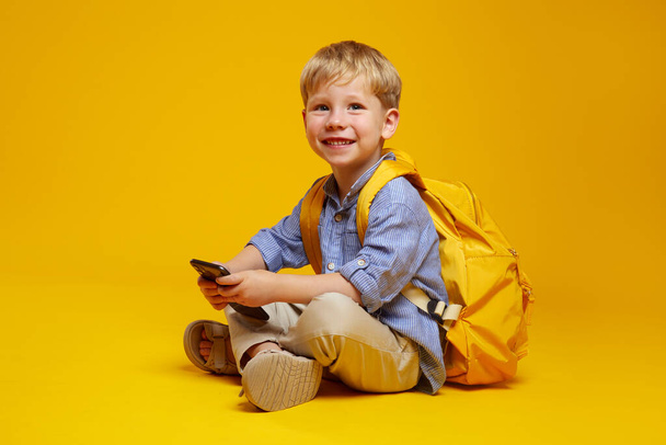 Radosny uczeń w niebieskiej koszuli z żółtym plecakiem trzymający telefon komórkowy w rękach, uśmiechający się do kamery siedząc na podłodze ze skrzyżowanymi nogami, odizolowany od pomarańczowego tła. - Zdjęcie, obraz
