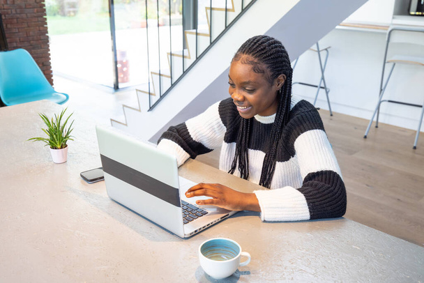 Αιχμαλωτίζοντας την ουσία της απομακρυσμένης εργασίας, μια νεαρή γυναίκα με πλεγμένα μαλλιά χαμογελά έντονα καθώς πληκτρολογεί μακριά στο laptop της. Το σύγχρονο σπίτι ρύθμιση, πλήρης, με κομψό εσωτερικό και φυσικό φως - Φωτογραφία, εικόνα