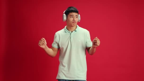 Oververheugd jonge Aziatische man in casual kleding dansen van vreugde tijdens het luisteren favoriete muziek tegen levendige rode studio achtergrond. Concept van menselijke emoties, mode en schoonheid, zelfexpressie, werk - Video
