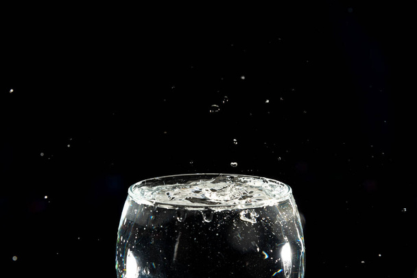 Spetterend water druppelt in een volle glazen beker. Splash Effect. Geïsoleerd op donkere achtergrond. - Foto, afbeelding