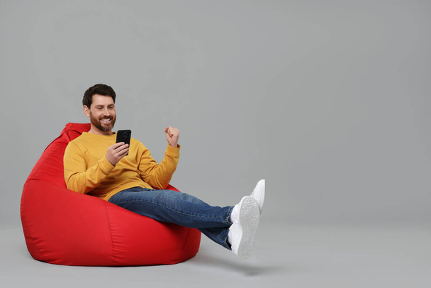 Hombre feliz con teléfono inteligente sentado en la bolsa de frijoles sobre fondo gris. Espacio para texto - Foto, imagen