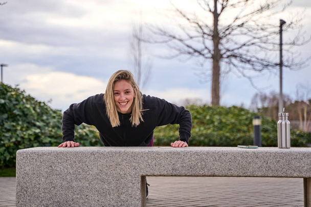 jonge vrouw doet biceps push-ups op een straat bank in de straat - Foto, afbeelding