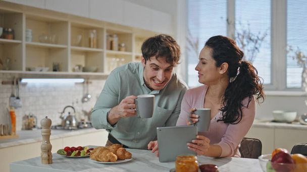 Ζευγάρι αναζητούν δισκίο πίνοντας πρωινό καφέ μαζί στην άνετη κουζίνα από κοντά. Χαρούμενοι σύζυγοι γελάνε συζητώντας για online αγορά κατά τη διάρκεια του πρωινού στο σπίτι. Ευτυχισμένο ζευγάρι με χρήση υπολογιστή καρτέλας. - Φωτογραφία, εικόνα