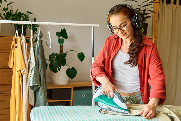 Glückliche Frau mittleren Alters in drahtlosen Kopfhörern, die Musik, Hörbücher oder Podcasts hört, während sie zu Hause gewaschene Wäsche auf dem Bügelbrett bügelt. Hausfrau-Alltag. Hauswirtschaftliches Konzept. - Foto, Bild