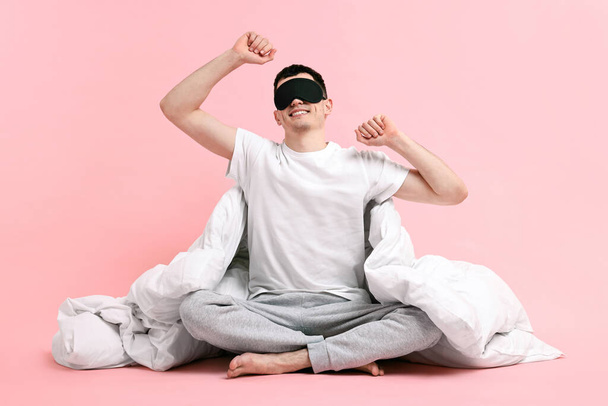 Happy man in pyjama with sleep mask and blanket on pink background - Фото, изображение