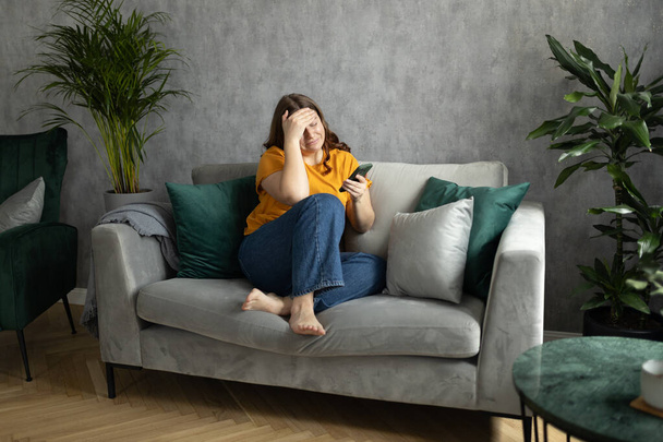 Portret pięknej młodej kobiety z depresyjnym wyrazem twarzy siedzącej na kanapie trzymającej telefon. Smutne kobiety po trzydziestce rozpaczliwie patrzące na ekran smartfona. - Zdjęcie, obraz
