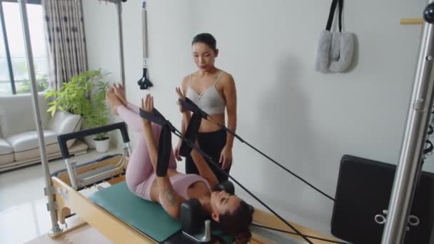 Weitschuss eines asiatischen Trainers hilft einer jungen Sportlerin bei Stretching-Übungen an Fitnessgeräten in Innenräumen - Filmmaterial, Video