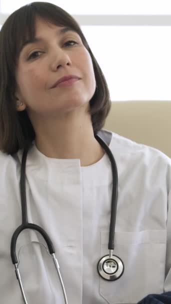 Giovane medico donna fiduciosa che scrive e sorride guardando la fotocamera con stetoscopio intorno al collo seduta sulla sedia in clinica - Filmati, video