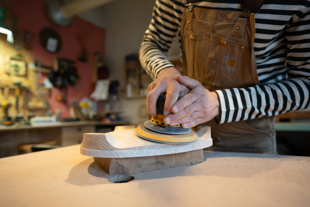 Nahaufnahme des Holzschleifens mit Orbitalschleifer in der Werkstatt. Tischler poliert Holzsitz eines zukünftigen Stuhls mit Elektroschleifer. Tischlerei. Möbelproduktion - Foto, Bild