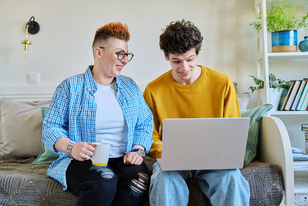 Middle-aged mother and son 19-20 years old sitting together on couch at home looking at laptop screen talking. Lifestyle family communication parenthood tenderness positive relationship two generation - 写真・画像