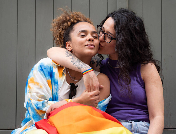 Las mujeres amantes de las lesbianas están abrazando y besándose. Llevan una bandera de arco iris y una pulsera para apoyar a la comunidad LGBTQ. - Foto, imagen