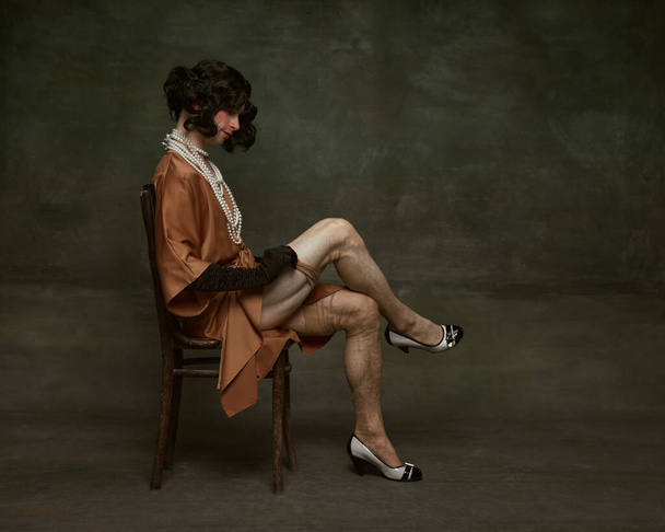 Ritratto laterale di giovane in abito seduto su una sedia di legno e calze aderente sullo sfondo vintage studio. Concetto di auto-espressione, confronti di epoche, libertà, diritti umani. - Foto, immagini