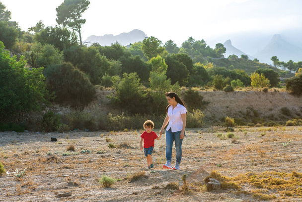 A woman and her young child holding hands while walking on a sunny nature trail with mountains - Photo, Image
