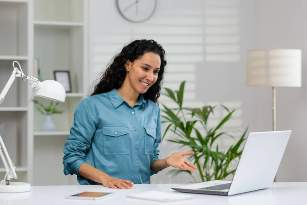 Friendly woman smiling and gesturing at the camera during a video call with a bright home office background. - Foto, Imagen
