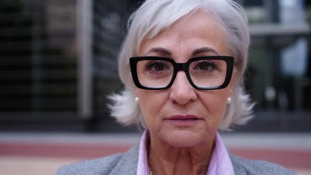 Portrait of mature Caucasian business woman with glasses and gray hair in formal suit smiling looking to the camera outdoor. Female senior looks serious and confident front of work place building - Materiał filmowy, wideo