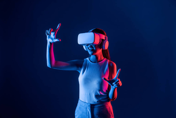 Vrouwelijke stand omgeven door neon licht dragen VR-headset aansluiten metaverse, futuristische cyberspace community technologie, verspreid zowel de hand index en duim vinger interactie virtueel object. Hallucinatie. - Foto, afbeelding